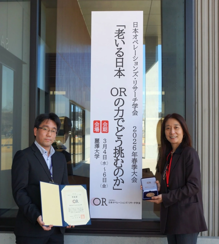 日本オペレーションズ・リサーチ学会の2026年春季大会のバナーの前で、男性が賞状を、女性がメダルを持って立っています