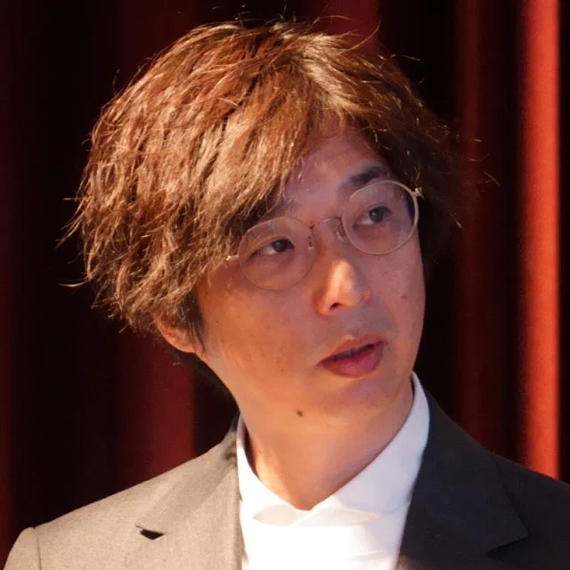 眼鏡をかけた男性が、茶色の髪とスーツ姿で、真剣な表情で何かを見つめている様子