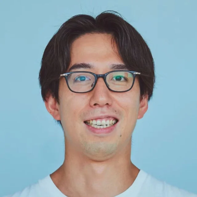 明るい水色の背景を背に、眼鏡をかけた若い男性が笑顔で写っているポートレート