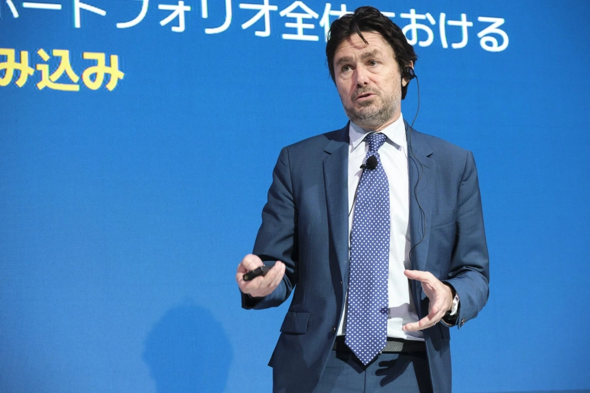 青い背景の前でスーツを着た男性がプレゼンテーションを行っています。彼は手にリモコンを持ち、マイクを装着して話しており、ビジネス会議やセミナーの様子を捉えています。
