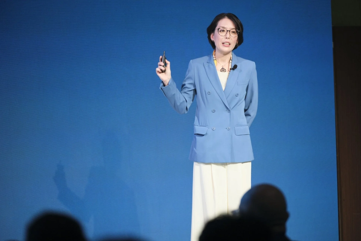 青い背景のステージで、青いブレザーを着た女性がプレゼンテーションを行っています。手にはクリッカーを持ち、聴衆に向かって話している様子です。