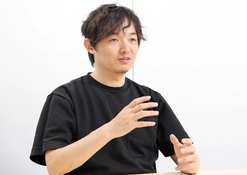 黒いTシャツを着たアジア人男性が、白い背景の前で座り、手を使って何かを説明している様子の画像です。カジュアルな雰囲気の会話やインタビューの場面を捉えています。