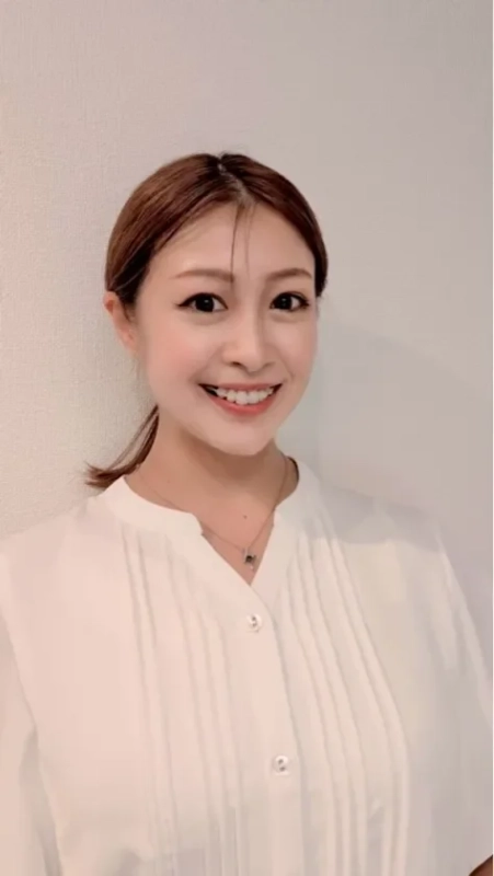 白いプリーツブラウスを着た若い女性が、カメラに向かって明るく微笑んでいます。髪はポニーテールにまとめられ、シンプルなネックレスを身につけています。