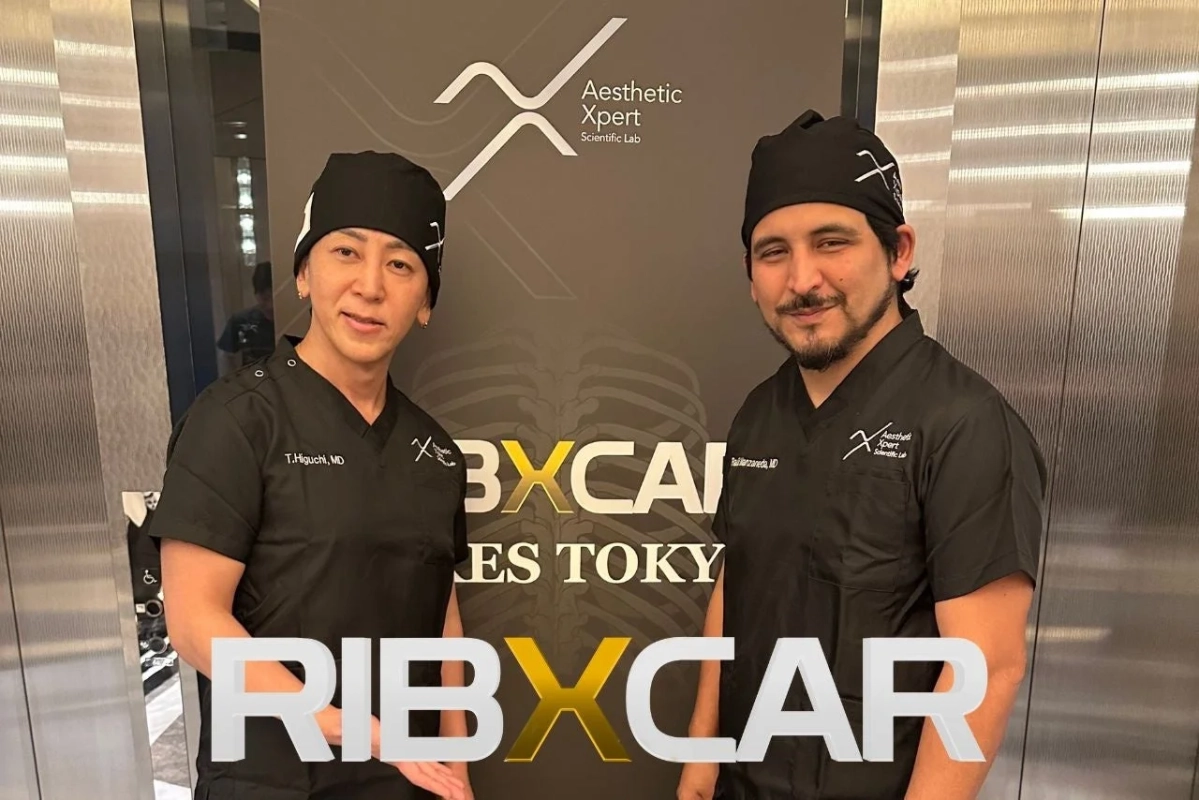 黒いスクラブを着た2人の男性医師が、Aesthetic XpertとRIBXCARのロゴが描かれたパネルの前で立っています。